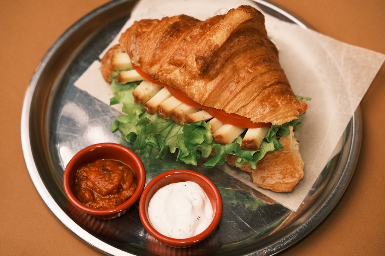 Croissant sandwich with cheese, lettuce, and tomato, served with red bowls of sauce and creamy dip.